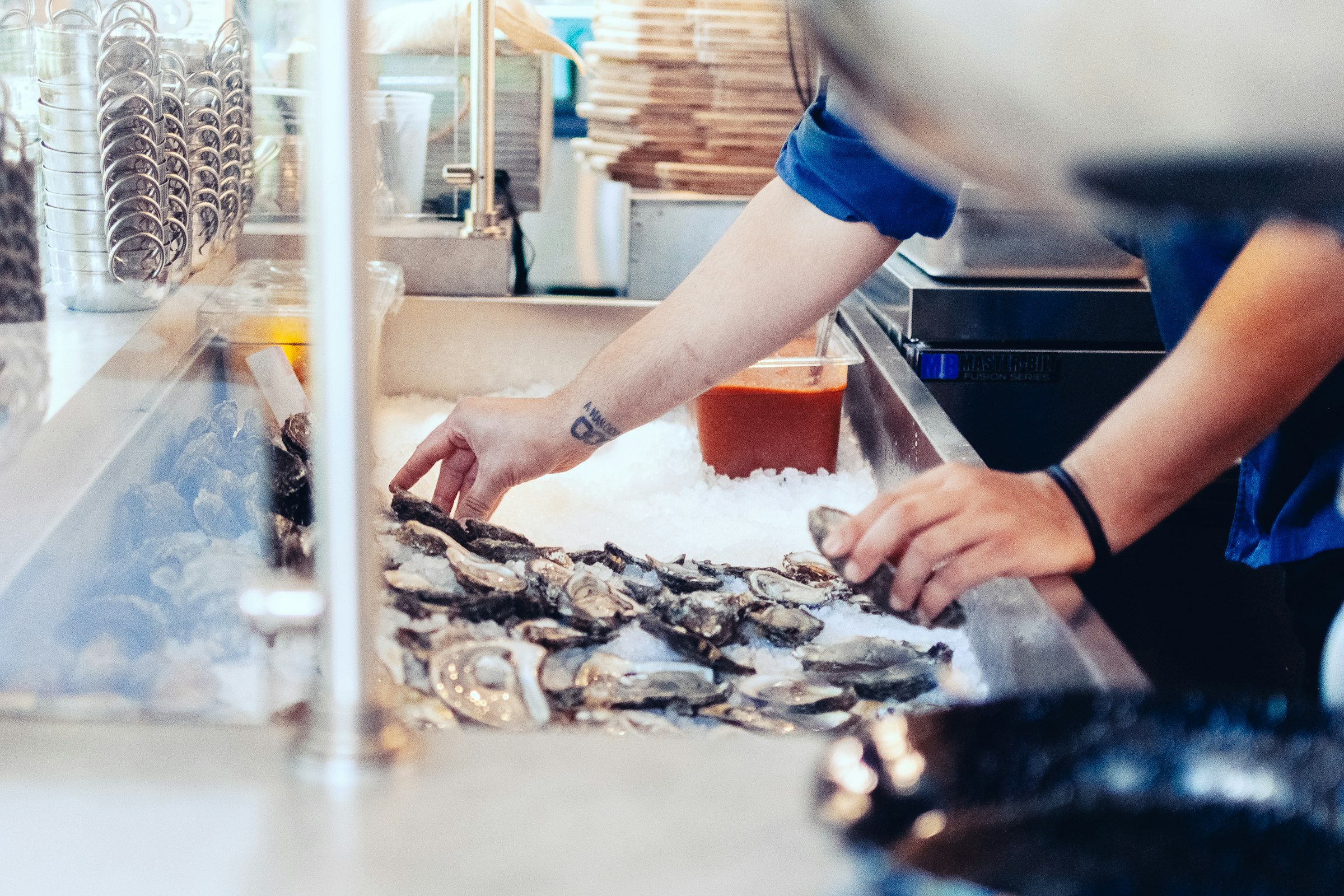 Restaurant worker's hands selecting oysters from a crushed-ice seafood display behind the counter
